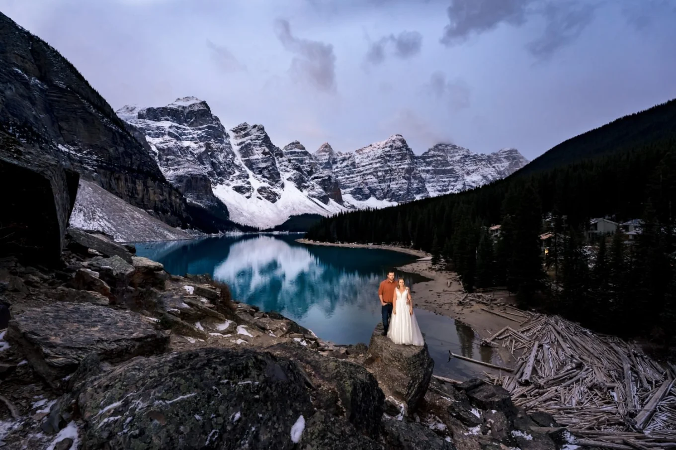 Incredible Banff Wedding Portrait at Moraine Lake of newlyweds standing at the stone. Behind them turquoise lake and Canadian Rocky Mountains covered with snow.