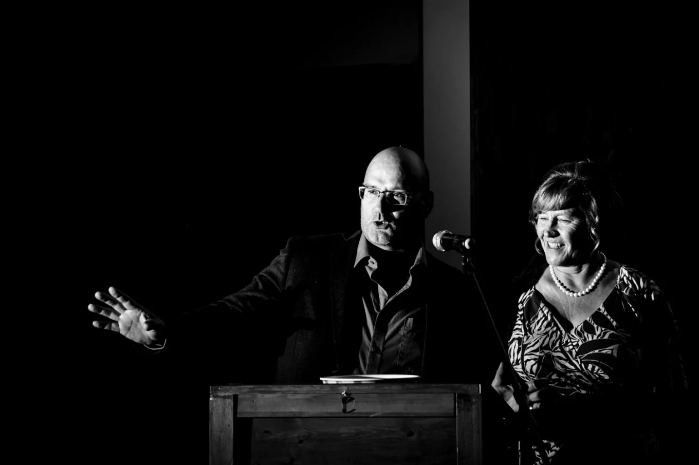 Wedding speech caltured by Calgary Wedding Photographer. Man talks to the microphone and lift his hand. Next to him his wife .
