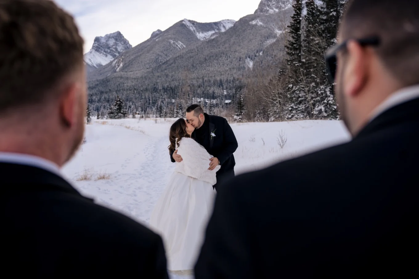 Bride and groom share a kiss while guests are watching them. Photo taken in Banff with the Rocky Mountains on the background.
