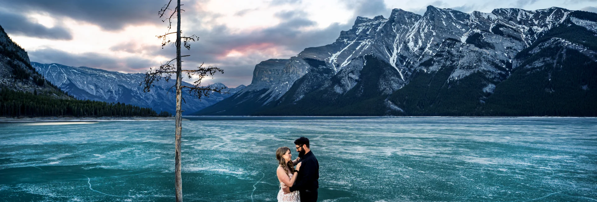 Engaged couple facing each other by the frozen Minnewanka Lake.