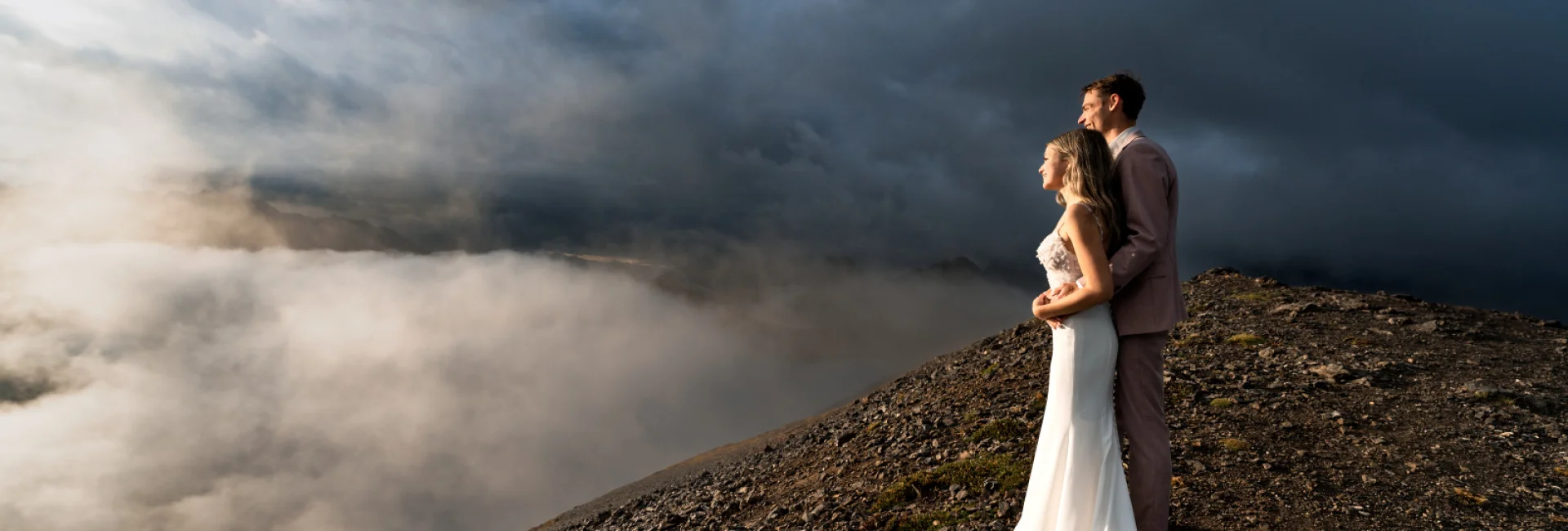 A blissful wedding couple stands atop a mountain, surrounded by ethereal clouds, embracing their love amidst breathtaking scenery in Banff.
