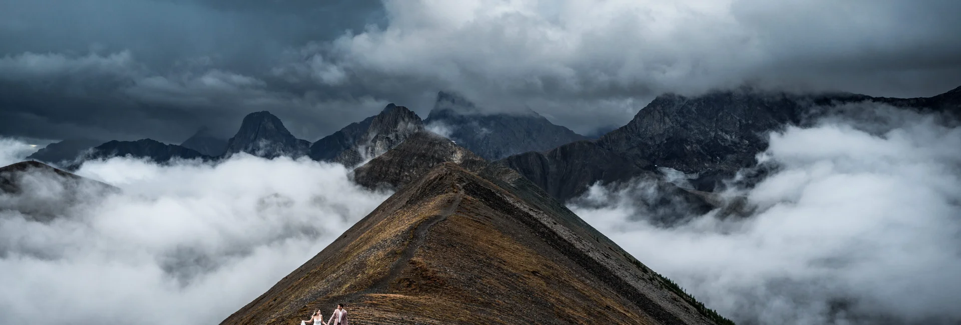 A couple in wedding attire stands on a mountain peak, overlooking a breathtaking view in Banff.