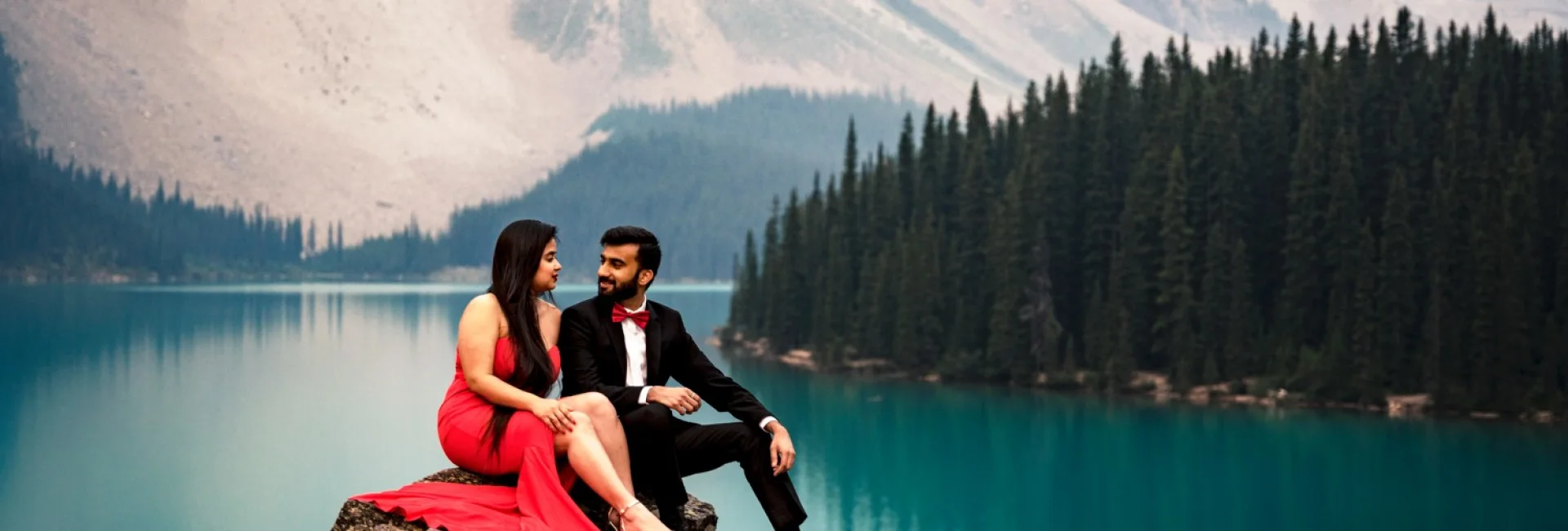 Engaged people sitting at the giant rock and look at each other during Moraine Lake engagement photo session with 4Eyes Photography Canada.