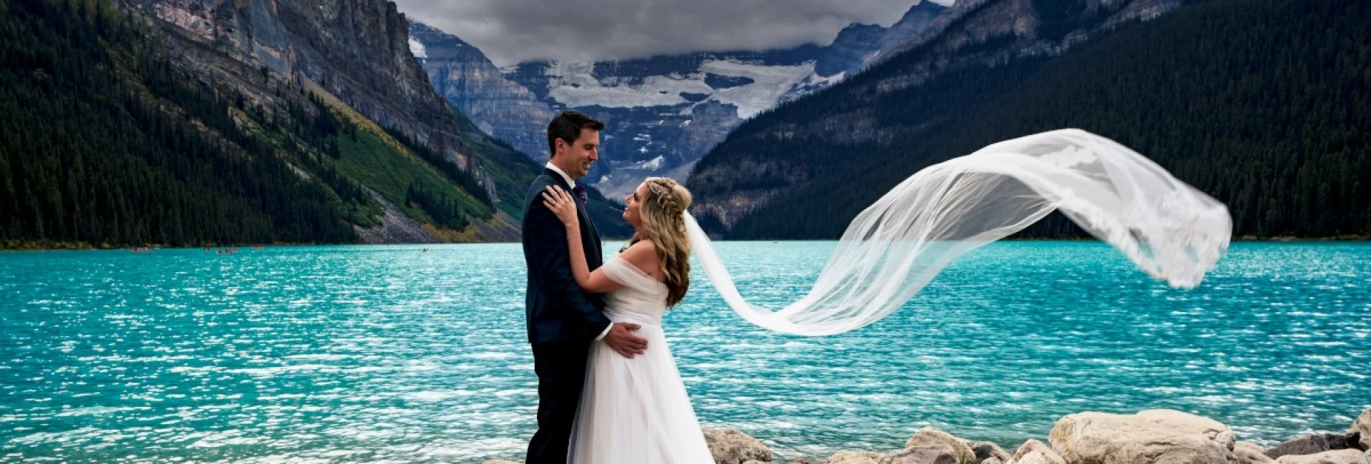 Newlyweds look deeply at each other's eyes. They are posing to Banff Wedding Photographer at Lake Louise. Bride's veil is blown by strong wind.