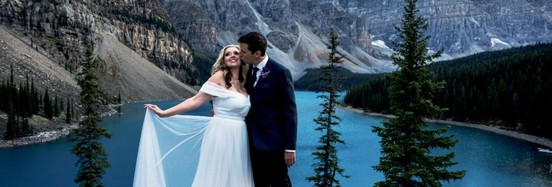Newlyweds standing at the giant rock at Moraine Lake. They are surrounded by the Canadian Rockies. She holds her dress and he kisses her cheek.