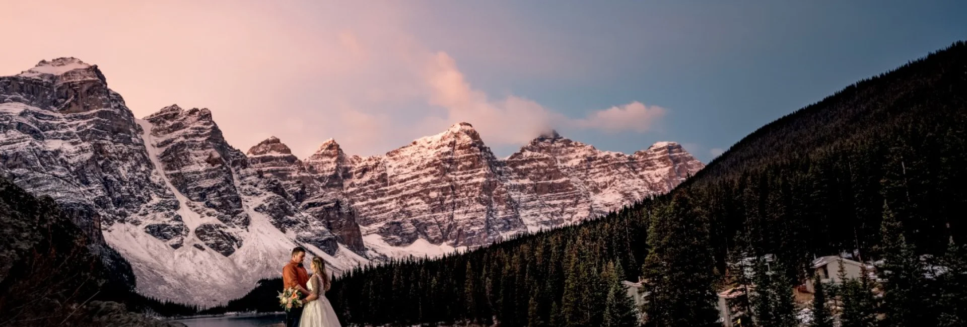 Newlyweds stand at the giant rock at Moraine Lake. They are surrounded by huge Canadian Rockies, Photo was taken during beautiful sunrise by Calgary Wedding Photographer.