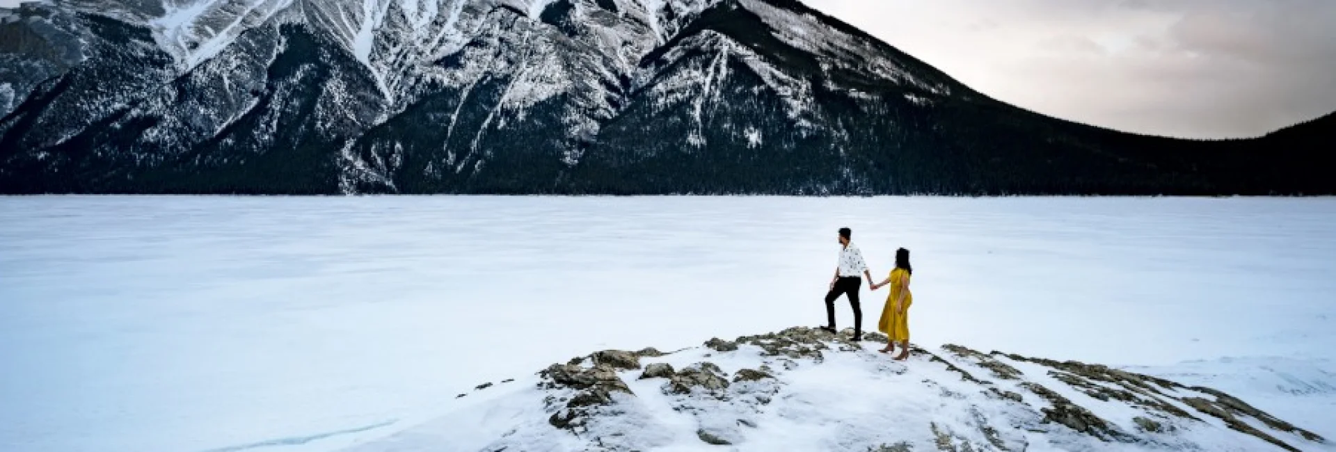 Incredible engagement photography taken by Calgary Wedding Photographer at Minnewanka Lake in wintertime. Couple standing at the giant rock and admire the beauty of the mountains.
