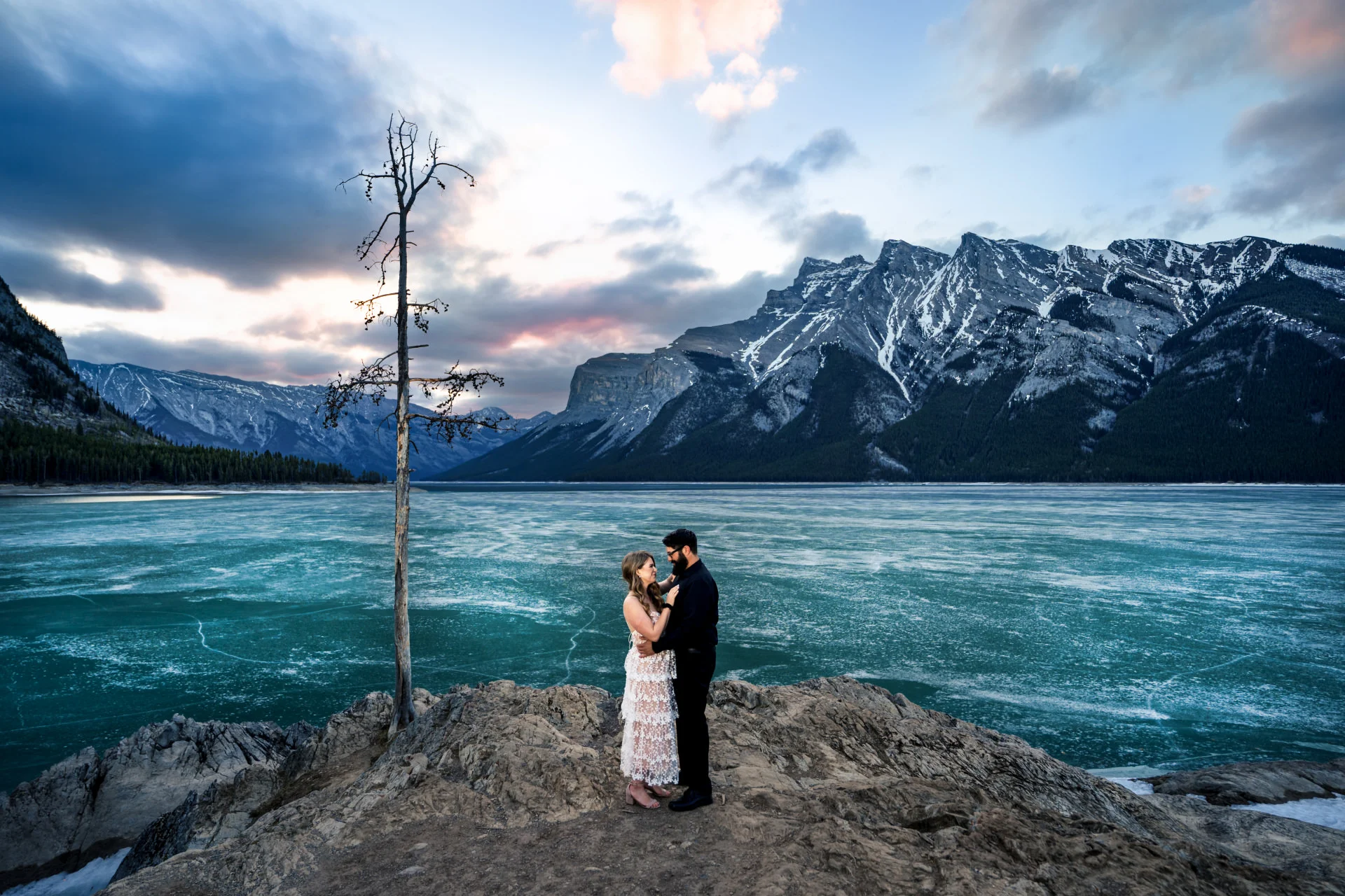 Engaged couple facing each other by the frozen Minnewanka Lake.