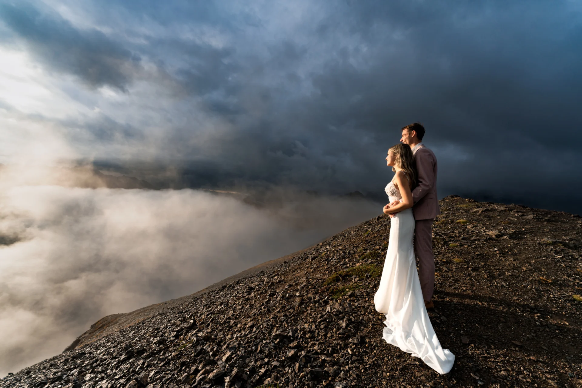 A blissful wedding couple stands atop a mountain, surrounded by ethereal clouds, embracing their love amidst breathtaking scenery in Banff.