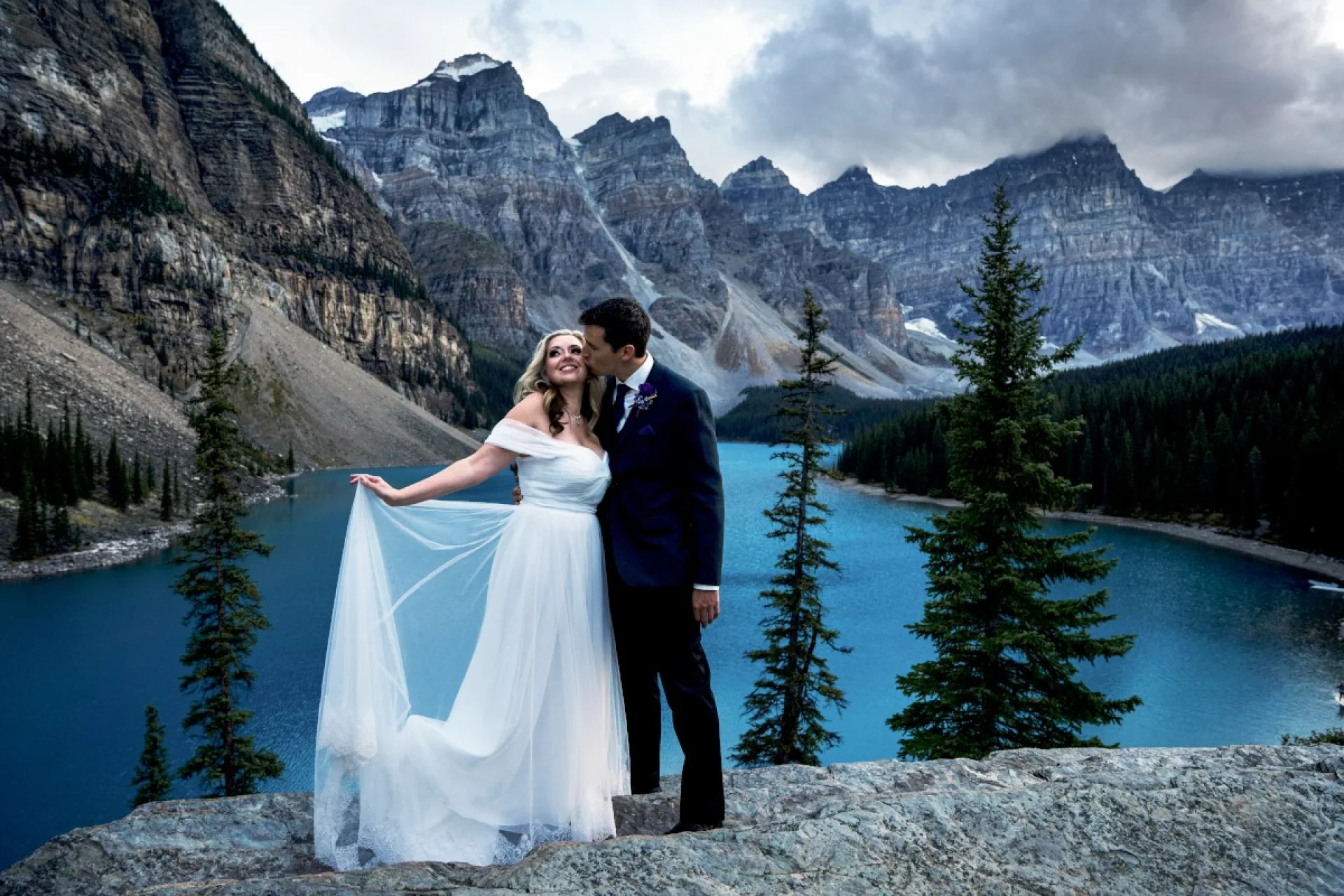Newlyweds standing at the giant rock at Moraine Lake. They are surrounded by the Canadian Rockies. She holds her dress and he kisses her cheek.