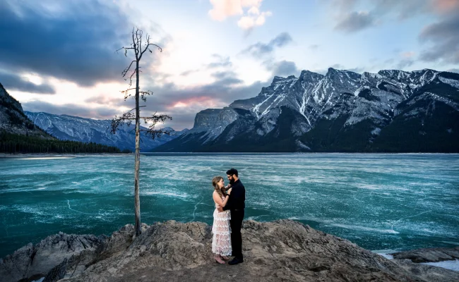 Engaged couple facing each other by the frozen Minnewanka Lake.