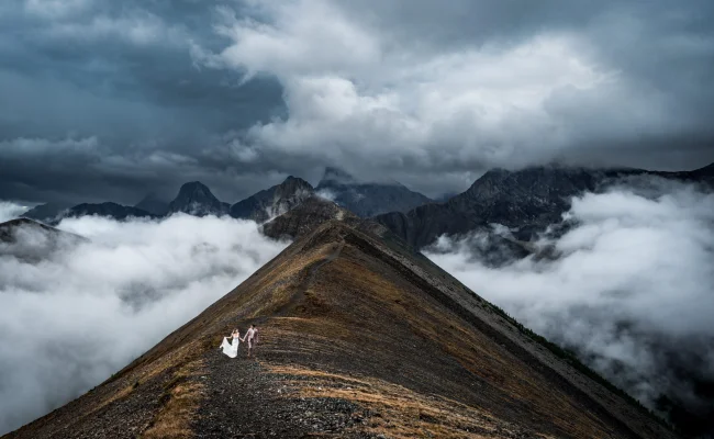 A couple in wedding attire stands on a mountain peak, overlooking a breathtaking view in Banff.