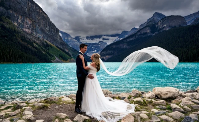 Newlyweds look deeply at each other's eyes. They are posing to Banff Wedding Photographer at Lake Louise. Bride's veil is blown by strong wind.