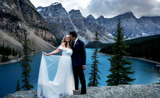 Newlyweds standing at the giant rock at Moraine Lake. They are surrounded by the Canadian Rockies. She holds her dress and he kisses her cheek.