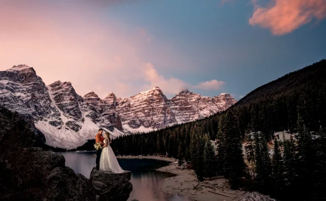 Newlyweds stand at the giant rock at Moraine Lake. They are surrounded by huge Canadian Rockies, Photo was taken during beautiful sunrise by Calgary Wedding Photographer.