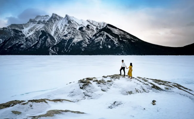 Incredible engagement photography taken by Calgary Wedding Photographer at Minnewanka Lake in wintertime. Couple standing at the giant rock and admire the beauty of the mountains.