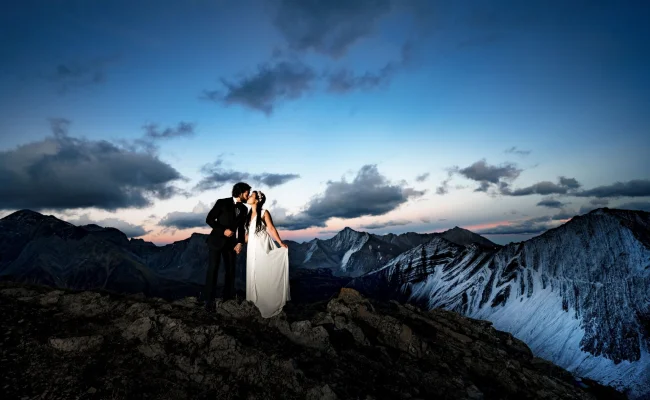 Newlyweds on the of the mountain in Banff posing to Banff Wedding Photographer. Against them stunning view of the Canadian Rockies.