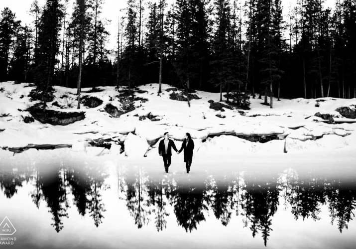 Engaged couple walk at the frozen lake looking at each other an holding hands. Behind them forest which is reflected on the bottom.