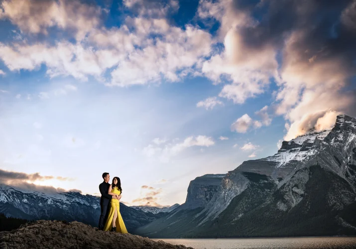 A man hugging his fiancee in a yellow dress in the stunning location of Minnewanka Lake in Banff during amazing sunrise.