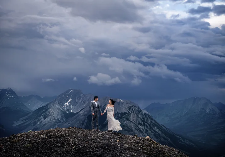 Dramatic Banff Wedding Photograph of the newlyweds holding their hands on the top of the mountain. Being them heave clouds and incredible view of the Canadian Rockies.