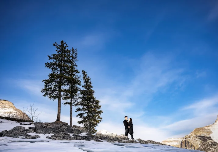 Tiny people in a hug up the hill. Next to them three trees and blue sky. Photograph taken in Banff by Calgary Wedding Photographer.