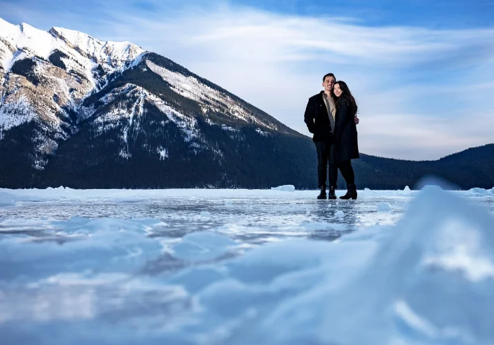 Young man and a woman in a hug at the frozen Minnewanka Lake. Behind them giant mountain.