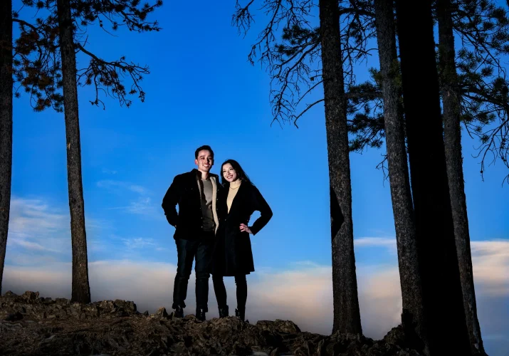 Handsome lovers posing for Calgary Wedding Photographer at Minnewanka Lake. They stand up the hill, between trees during sunset.