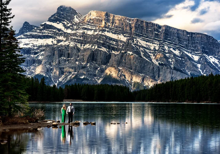 Woman in a green dress and her fiance hold their hands at the stunning Two Jack Lakes in Banff. In the background tremendous mountain.