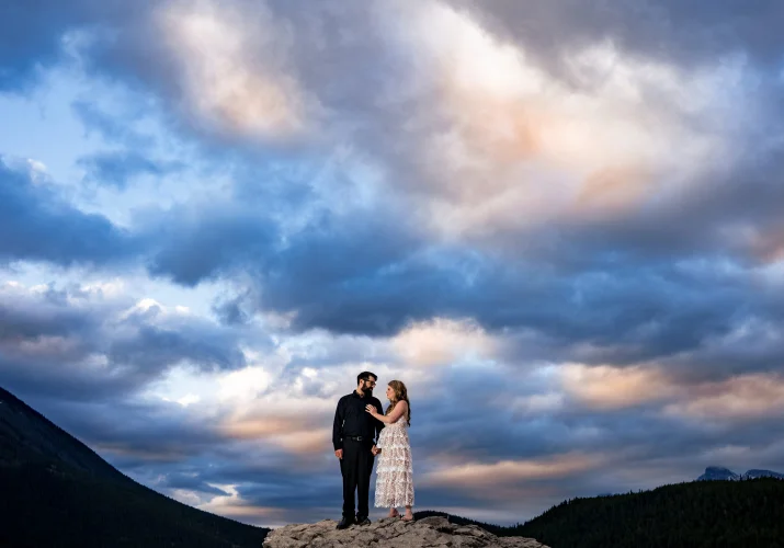 Woman in a white dress touches her fiance's chest and look deeply into his eyes. Above them dramatic sky.