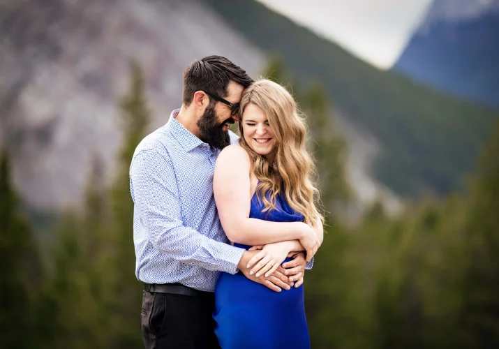 Man with long black beard whispers a secret into his fiancee's ear. They both laugh.