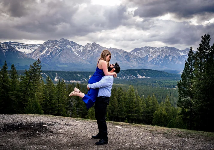 Gentleman lifts his fiancee. She wears a blue dress. They are in the mountains in Banff National Park.