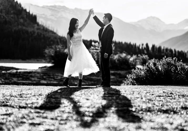 Creative photograph in black and white taken by Banff Wedding Photographer. Couple dance by the lake while they shadows are visible at the ground.