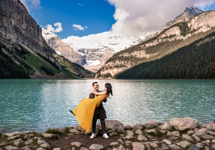 Man lifting his fiancee in a yellow dress at the stunning location of Lake Louise. Behind them lake and the Canadian Rockies.