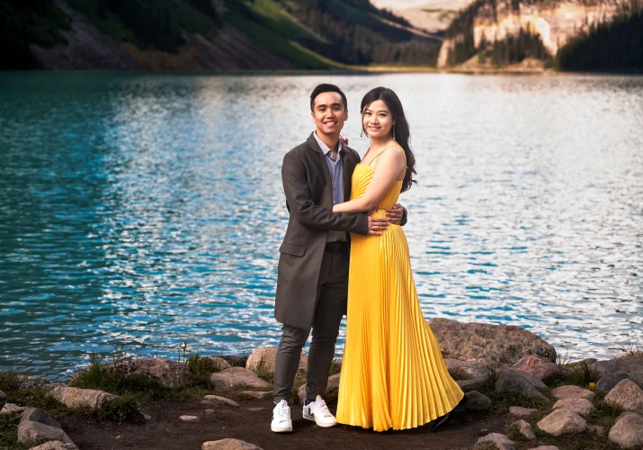 Engaged couple stand in ahead and look at the camera by the lake.