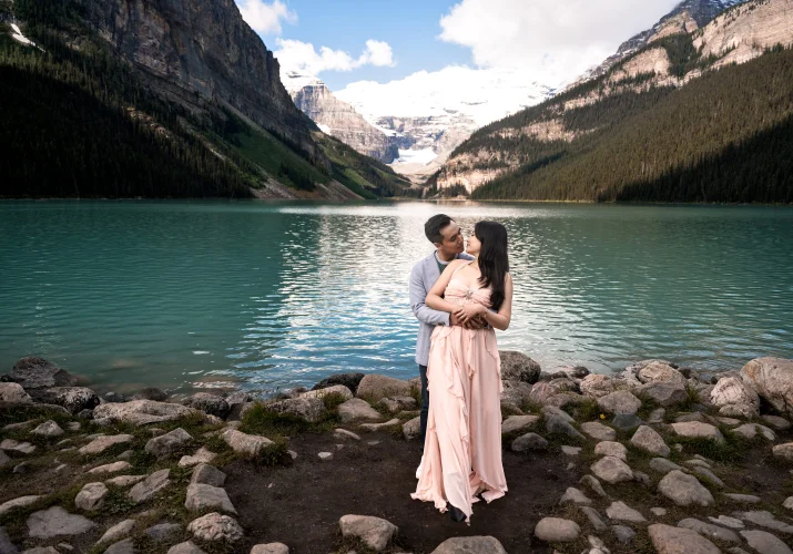 A man hug his fiancee from behind, they look at each other at the edge on Lake Louise.