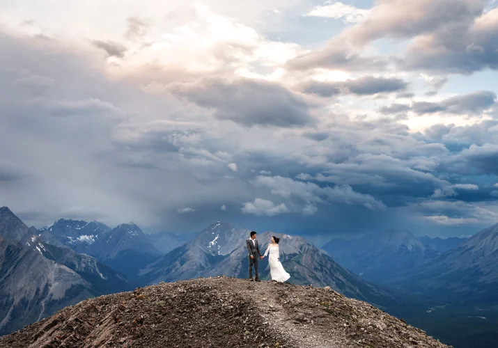 Tiny bride and groom at the ridge of the mountain siding sunset with the heavy clouds above them.