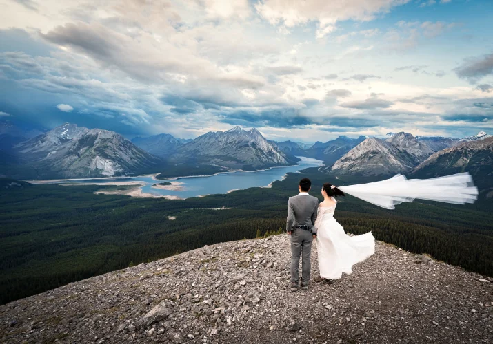 Bride and groom adore the stunning view of the Canadian Rockies and a blue lake from the peak.
