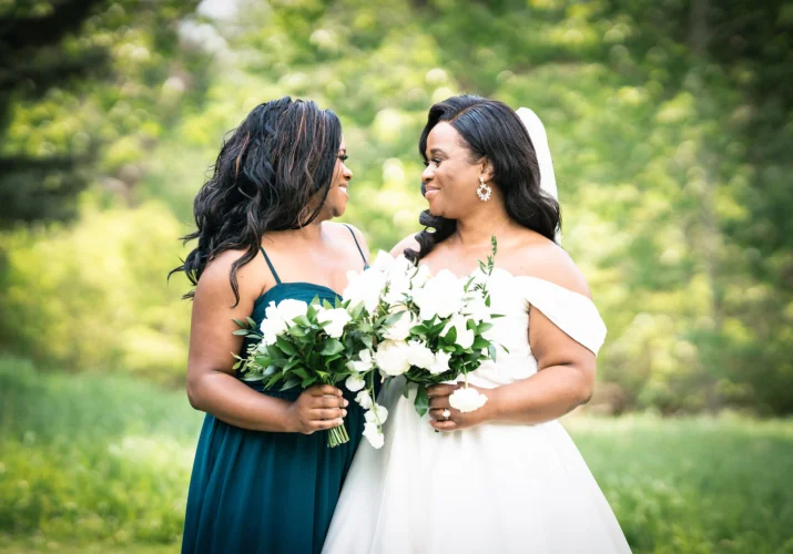 Portrait on the bride and maid of honour looking at each other. They hold white flowers in their hands.