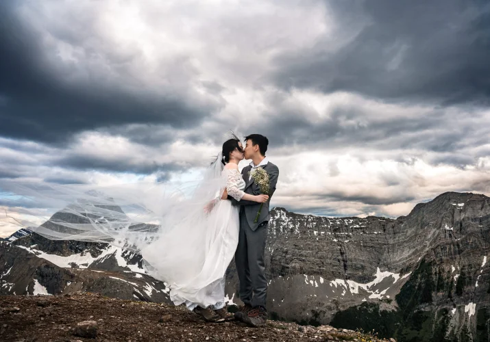 Bride in a long white dress and the groom share a kiss in Banff National Park. Above them heavy clouds during the sunset.
