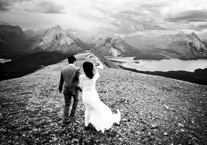 Newlyweds walk at the ridge of Tent Ridge with majestic view of the Rocky Mountains and the lake.