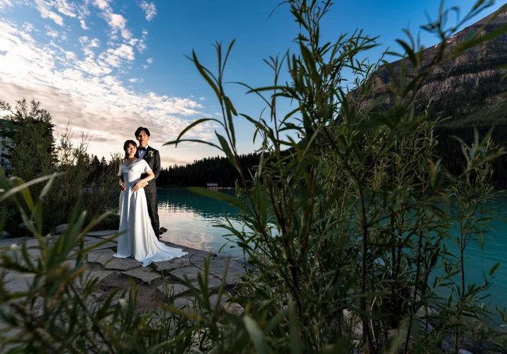 Groom hug his wife from behind. Photo taken through the bushes during the sunrise at Lake Louise.