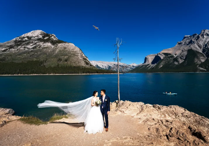 Stunning wedding portrait at Minnewanka Lake taken by Banff Wedding Photographer. Newlyweds standing next to each other a look at their eyes. Behind them Blue lake and Rocky Mountains.