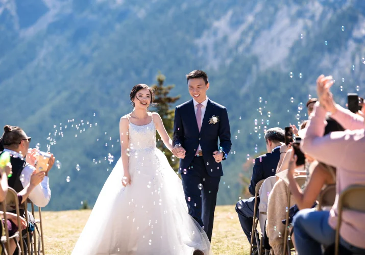 newlyweds walk out of the aisle holding their hands in Banff. All around them bubbles.