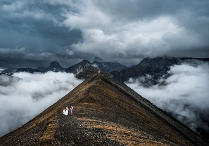 Fearless couple at the top of the mountain walking at the ridge in Banff. On the background stunning Rocky Mountains and heavy clouds.
