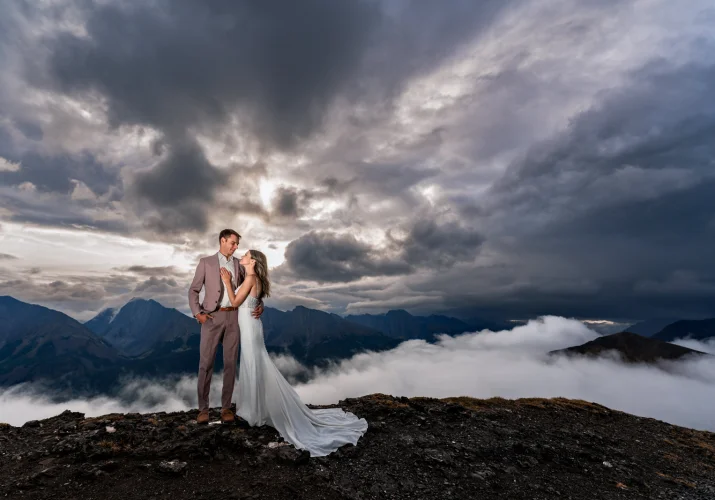 Bride hug the groom at the summit of the mountain in Banff. On the background huge Rocky Mountains and heavy clouds.