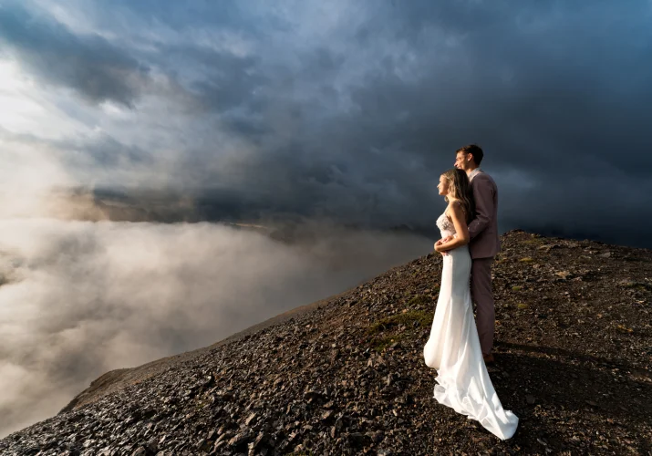 A blissful wedding couple stands atop a mountain, surrounded by ethereal clouds, embracing their love amidst breathtaking scenery in Banff.