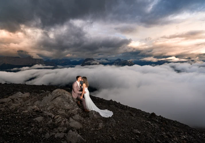 The bride kisses the groom who sits on the rock at the top on the mountain during coloreful sunset in Banff.
