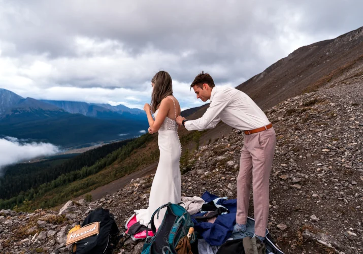 Groom helps the bride to zip her wedding dress before the wedding session in the mountains.