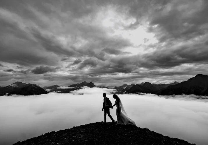A couple in wedding attire walks triumphantly on a mountain peak, embracing each other with joy and love.