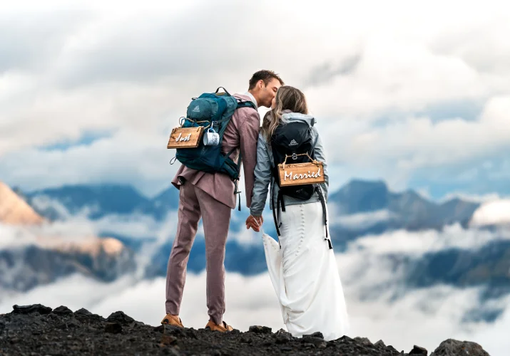 Bride and groom share a kiss at the summit of the Canadian Rockies wearing hiking backpacks.
